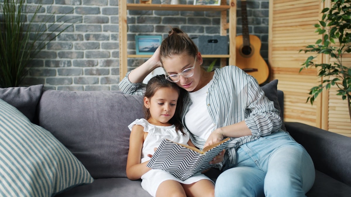 Mother and little daughter are reading book at home smiling relaxing on couch together enjoying children's literature. People, leisure and culture concept.